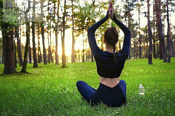 Schlanke schöne Frau in schwarzem eng anliegendem Trainingsanzug sitzt auf Gras im Wald in Yoga Pose