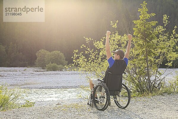 Happy and joyfull young man in wheelchair outside in nature on a sunny summer day with his hands raised up