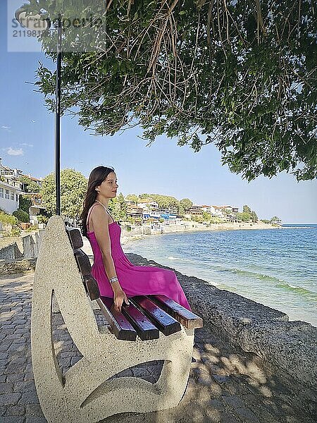 Schöne junge Frau in rosa Kleid  sitzt auf einer Bank und beobachtet das Meer und die Altstadt von Nessebar Küste. Sommerurlaub Reise Szene in Bulgarien