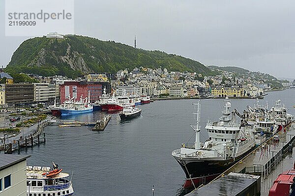 Harbour town with several ships  cloudy sky and adjacent buildings in front of a green hill  Alesund  Fylke  Norway  Europe