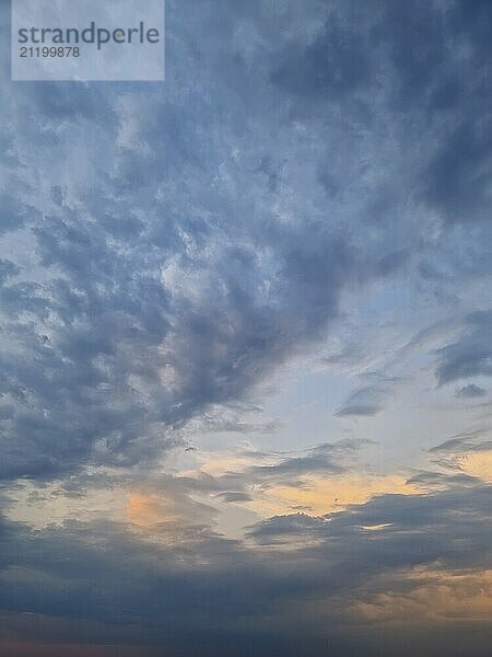 Eine weite Himmelsansicht mit dynamischen Wolkenformationen  Blautönen und zartem Orange am Horizont  die entweder einen Sonnenaufgang oder einen Sonnenuntergang suggerieren. Das Zusammenspiel von Licht und Wolkenlandschaft schafft eine heitere Atmosphäre