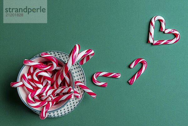 Coffee cup full of mini candy canes with red-white stripes  and a few on the green background in a heart shape. Christmas traditional sweets