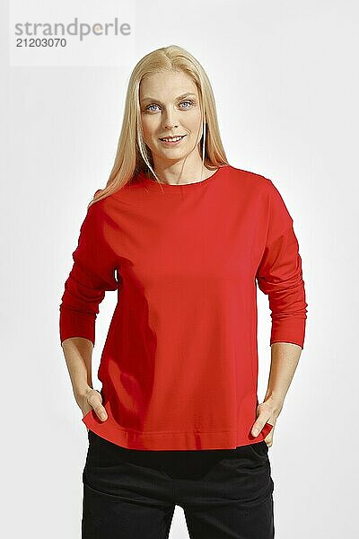 A smiling woman with long blonde hair poses confidently with her hands on her hips  wearing a vibrant red top. The background is plain white  emphasizing her cheerful demeanor and stylish outfit