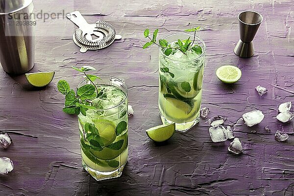 Mojito Cocktail  zwei Gläser. Sommerliches Kaltgetränk mit Limette  frischer Minze und Eis. Kühles Getränk auf einem lila Hintergrund mit Mischzubehör  Food Fotografie