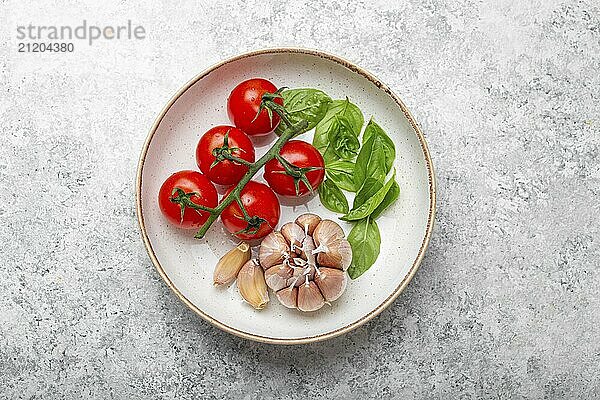 Italienisches Essen Kochen Konzept mit Kirschtomaten  frisches grünes Basilikum und Knoblauchzehen Draufsicht auf Teller auf weißem Stein Hintergrund  Zutaten für die Zubereitung von mediterranen Mahlzeit  Lebensmittel Fotografie