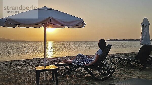 Sorglose junge Frau liegt auf einer Sonnenliege am Strand und beobachtet die Morgendämmerung mit Blick auf das Meer. Schöne Szene am Meer  Sommerurlaub Entspannung und Urlaub genießen Konzept