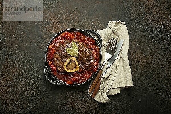 Traditionelles italienisches Gericht Ossobuco all Milanese aus geschnittener Kalbshaxe mit vegetarischer Tomatensoße  serviert in schwarzer Kasserolle von oben auf rustikalem braunem Hintergrund  Foodfotografie  food photography  food photography