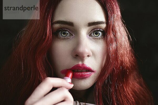 Portrait of crazy-looking depressed teen girl with red hair she is smearing red lipstick on her face  black background. social issues. social protest. The problem with raising teenagers