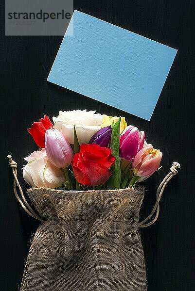 Women international day card with a bouquet of diverse flowers wrapped in a hessian bag and an unwritten blue message card on a black background