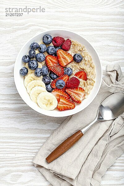 Haferflockenbrei mit Obst und Beeren in der Schüssel mit Löffel auf weißem Holzhintergrund Tischansicht  hausgemachte gesundes Frühstück Müsli mit Erdbeere  Banane  Heidelbeere  Himbeere. Platz für Text  Food Fotografie  food photography