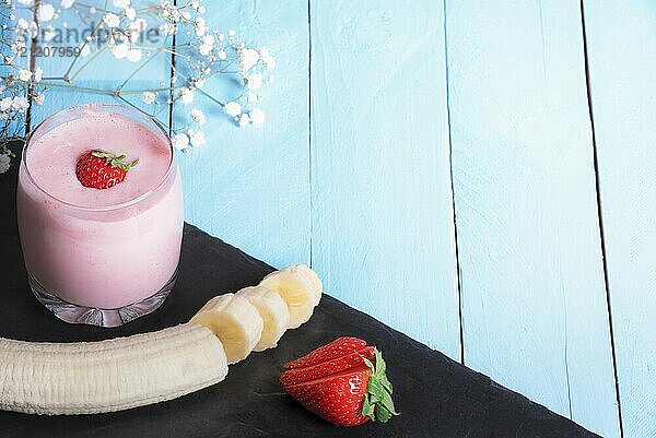 Healthy eating concept with a glass of fresh strawberry and banana smoothie  sliced fruits on a black plate  on a blue background  in sunlight