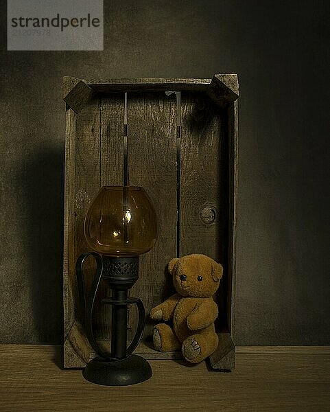 Still life with old teddy bear and a golden light in an old wooden box on a table with dark background