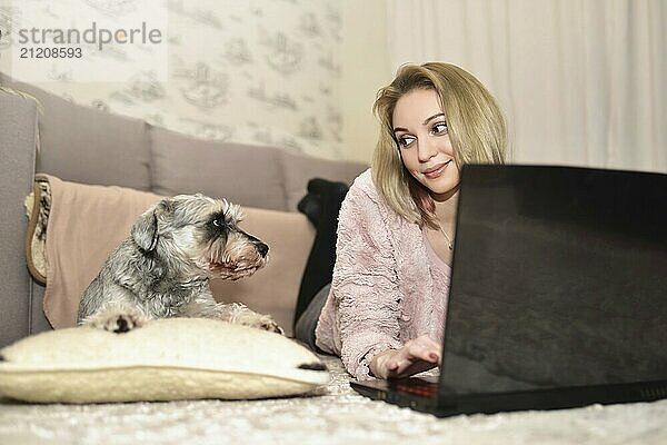 Young Lady is lying on the carpet at home with a laptop and looking at her cute gray dog who is asking for attention