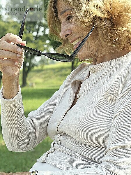 Serene senior woman lies in lush green grass in a peaceful park. Basking in the warm summer sun. Holding her sunglasses with a content smile. Embodying freedom and relaxation