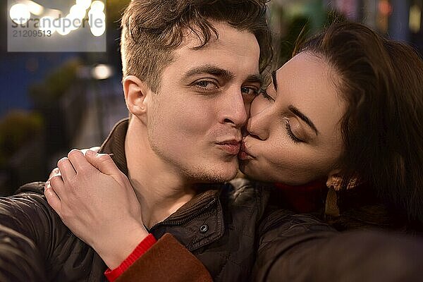 A young trendy couple in love walks down a night street and takes selfies. The girl is kissing her boyfriend