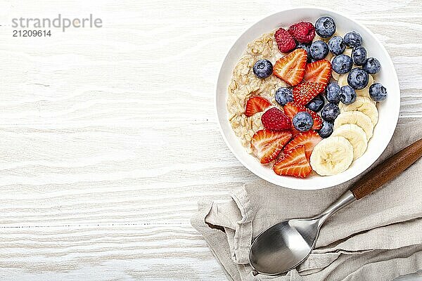 Haferflockenbrei mit Obst und Beeren in der Schüssel mit Löffel auf weißem Holzhintergrund Tischansicht  hausgemachte gesundes Frühstück Müsli mit Erdbeere  Banane  Heidelbeere  Himbeere. Platz für Text  Food Fotografie  food photography