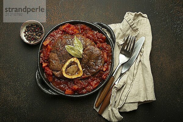 Traditionelles italienisches Gericht Ossobuco all Milanese aus geschnittener Kalbshaxe mit vegetarischer Tomatensoße  serviert in schwarzer Kasserolle von oben auf rustikalem braunem Hintergrund  Foodfotografie