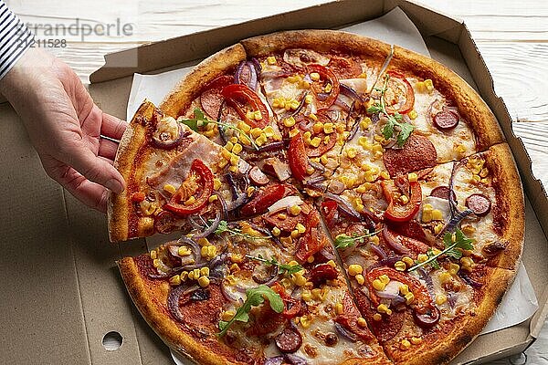 Human caucasian hand takes slice of large pizza from carton box on white wooden table