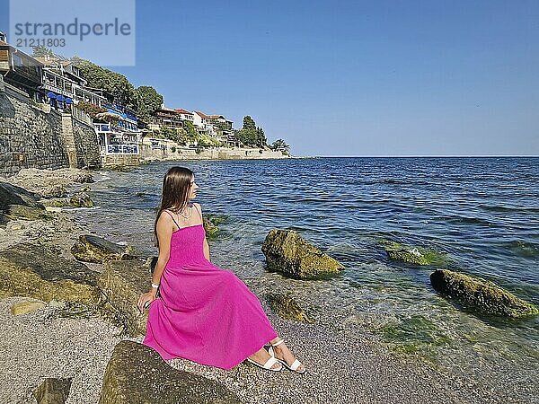 Schöne junge Frau in rosa Kleid  sitzt auf einem Felsen am Strand und beobachtet die Sehenswürdigkeiten der Altstadt von Nessebar in Bulgarien. Schöne alte Häuser an der Küste. Sommerurlaub Reise Szene