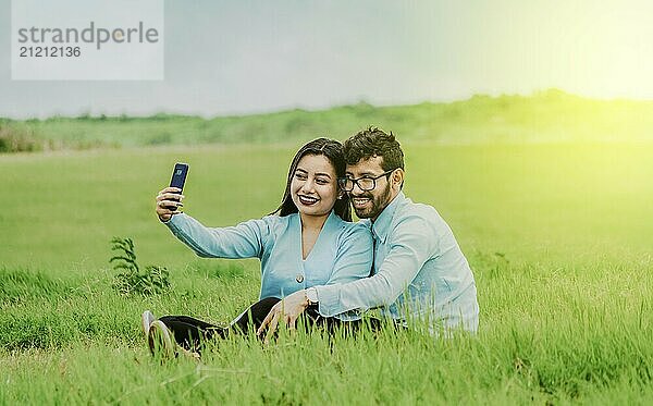 Smiling young couple in love taking a selfie sitting in the field. Happy Couple in love taking a selfie sitting in a beautiful field