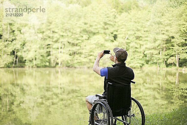 Happy and young disabled or handicapped man sitting on a wheelchair in nature. Photographing landscape with smartphone