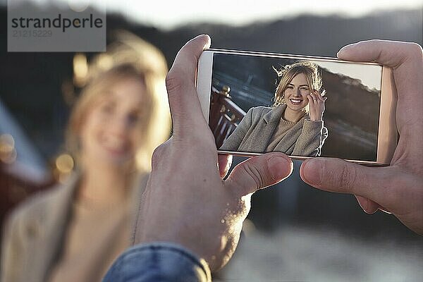 A man make a portrait on the phone of happy smiling woman standing on the bridge on sunny spring or autumn day outdoor