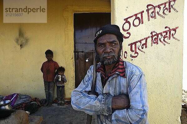 Porträt eines Mannes und von Kindern  die zum Stamm der Birhor gehören  Jharkhand  Indien. Sie stehen vor dem Einzimmerhaus  das die indische Regierung für sie gebaut hat. Der Name des Eigentümers steht in roter Schrift an der Wand. Früher waren die Birhor Nomaden  sie lebten vom Jagen und Sammeln. Nach der Unabhängigkeit Indiens im Jahr 1947 versuchte die Regierung  sie zu besiedeln  indem sie ihnen Land und Ochsen schenkte. Doch für viele von ihnen ist dieser Integrationsversuch gescheitert. Sie leben heute in großer Armut