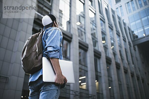 Outdoor portrait of handsome young male student with backpack standing at building background on the street while waiting his colleagues. Businessman wearing casual white shirt and spectacles. People