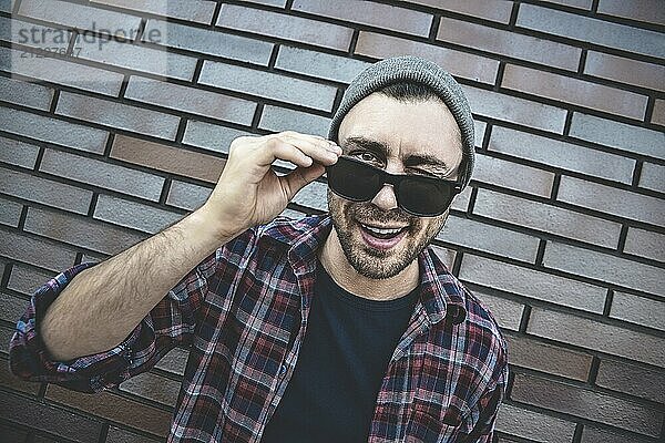 Young adult man wearing sunglasses standing over brown brick wall smiling with happy face winking at the camera