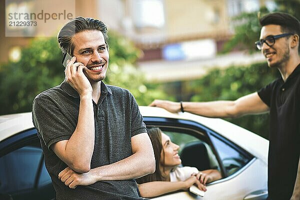 Handsome young caucasian man talking on the mobile phone and smiling while standing near the car with his friends chatting in the background