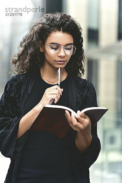 Charming Afro American beautiful happy female  making notes in notepad while walking outdoors on a city street. Education and business concept with space for text