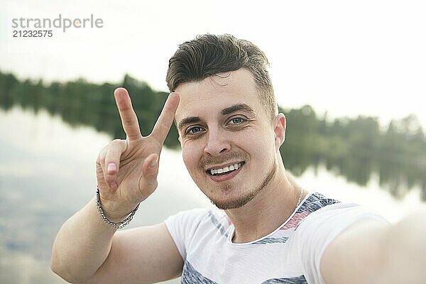 Portrait of Young handsome stylish smiling guy makes selfie against the lake. beautiful nature