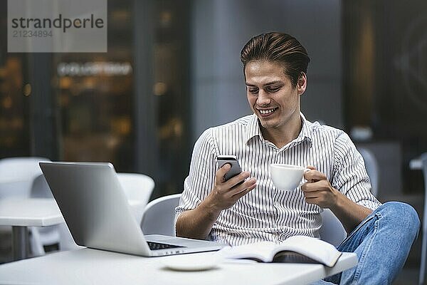 Handsome caucasian young man with phone working on laptop and smiling while enjoying coffee in cafe