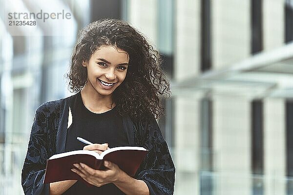 Charming Afro American beautiful happy female  making notes in notepad while walking outdoors on a city street. Education and business concept with space for text