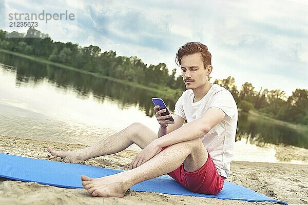 Man is sitting on the beach at lake and writing a sms on phone. good mood. beautiful view