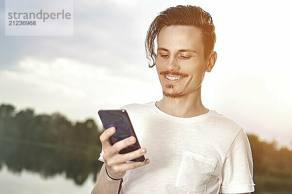 Young man walking near beautiful bay using phone and headphones for communicate with friends  good mood  beautiful view