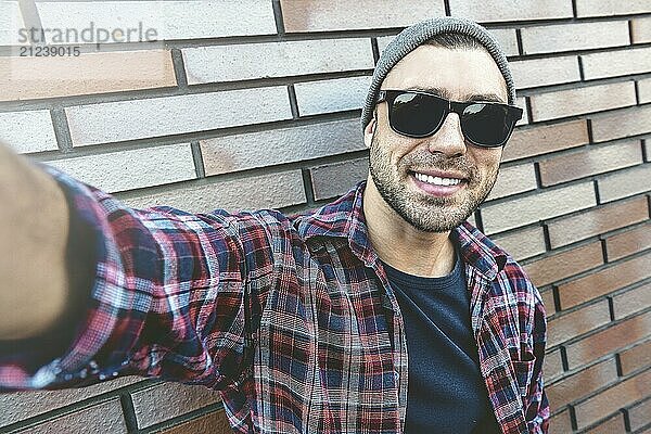 Portrait of stylish handsome young man with bristle standing outdoors and leaning on brick wall. Man wearing sunglasses and hat. Man making selfie