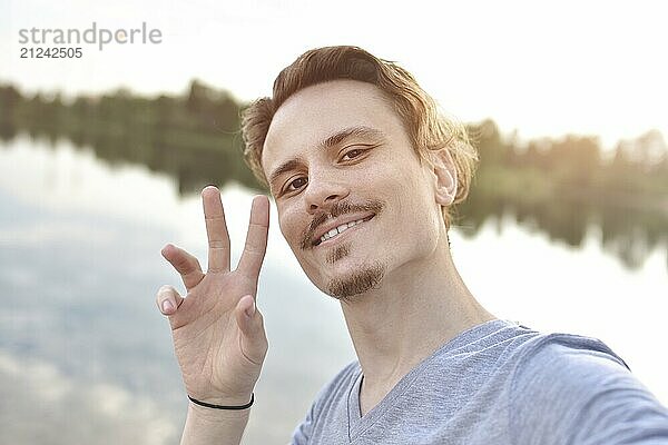 Portrait of Young handsome stylish smiling guy makes selfie against the lake. beautiful nature