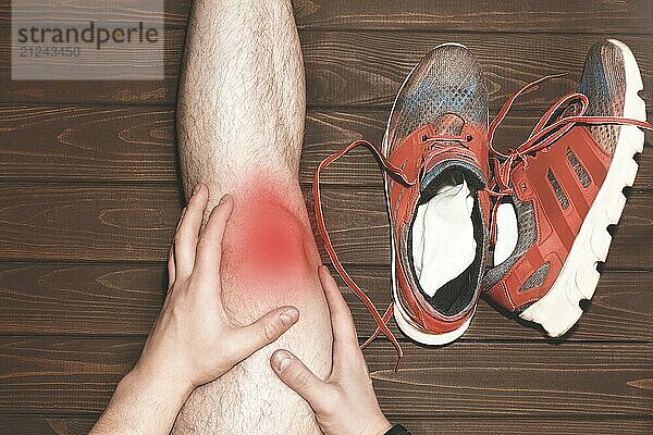 Young sport man with strong athletic legs holding knee with his hands in pain after suffering ligament injury during a running workout training on wooden background. red effect