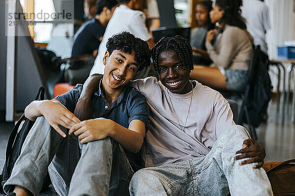 Portrait of happy teenage boy with arm around male friend sitting in cafeteria at school