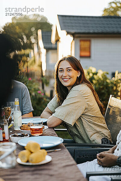 Portrait of smiling young woman sitting near dining table in back yard at dinner party