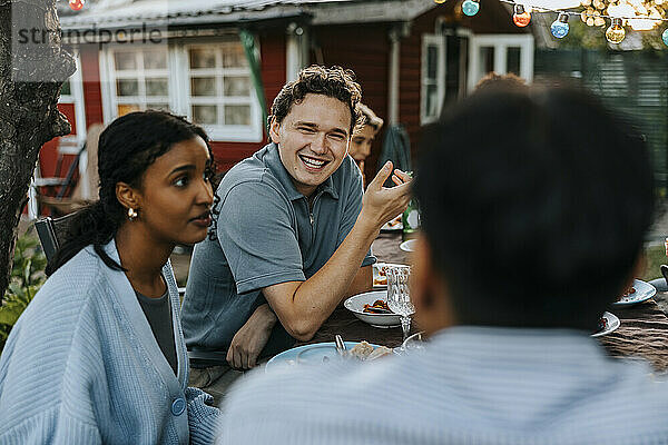 Happy young man talking with male friend while having dinner in back yard at social party