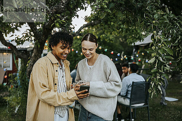 Smiling young man sharing smart phone with female friend while standing in back yard at social gathering