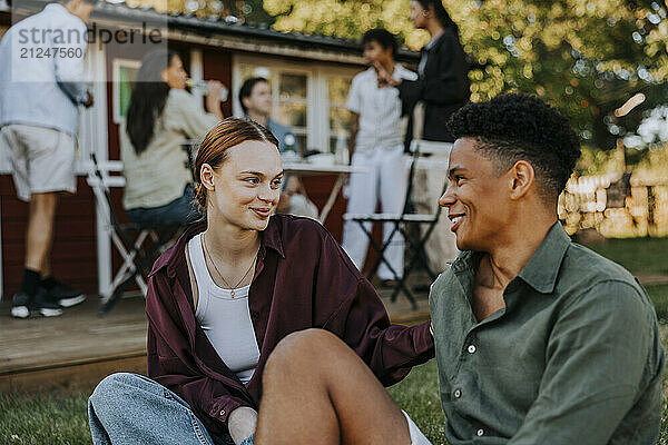 Smiling young woman talking with male friend while sitting on grass in back yard