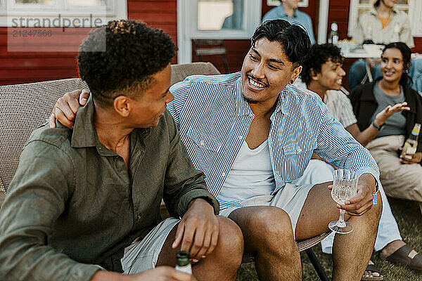 Smiling young man enjoying drink with male friend sitting on chair in back yard at dinner party