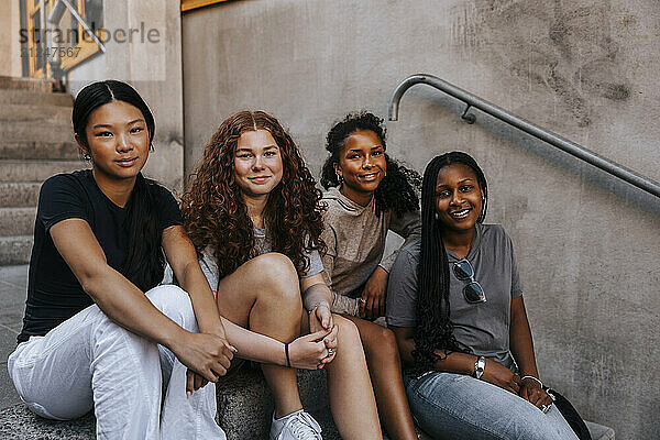 Portrait of smiling female friends sitting on stairs near wall of junior high school building