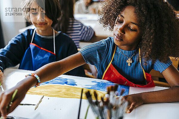 Focused curly hair girl painting on paper during art class at school