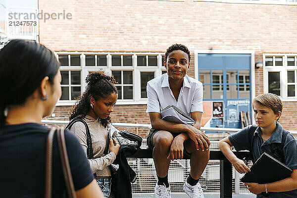 Teenage boy sitting on railing while talking with friends at balcony of junior high school