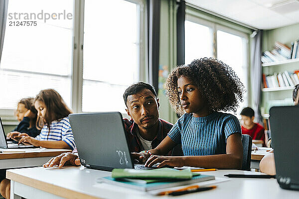 Male professor helping curly hair girl with studies on laptop in classroom at elementary school