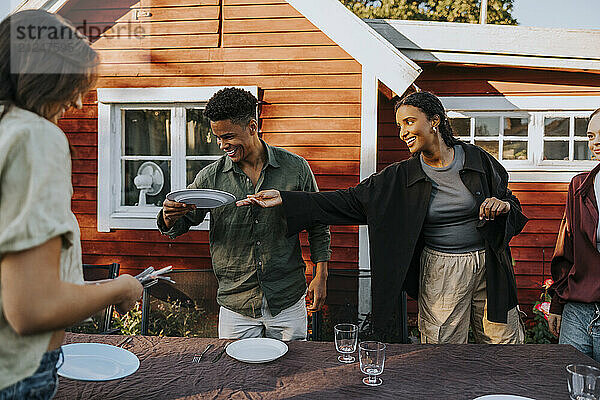 Smiling young woman passing plate to male friend while setting dinner table in back yard at social party